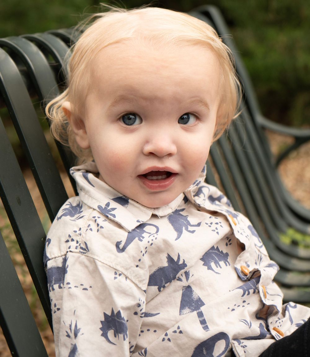 Adorable blonde toddler with blue eyes wearing a dinosaur print shirt, sitting on a green metal bench outdoors.