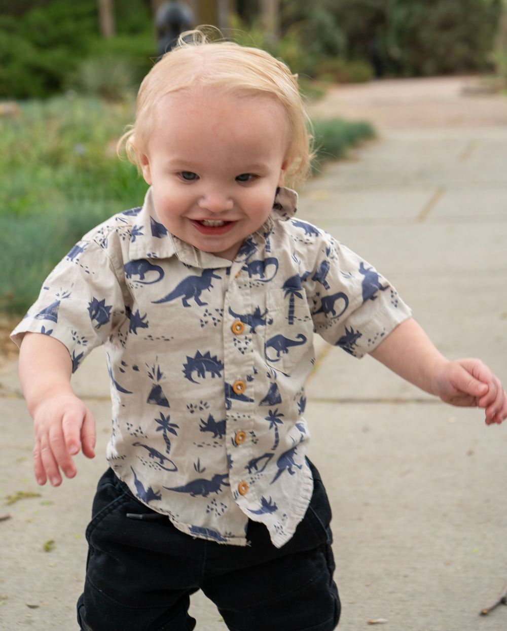 Smiling toddler boy in dinosaur print shirt running outdoors on a sunny day.