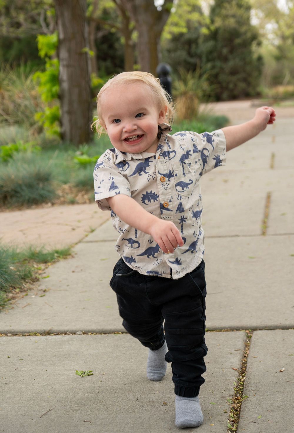 Happy toddler boy in dinosaur shirt and black pants running on a garden path outdoors.