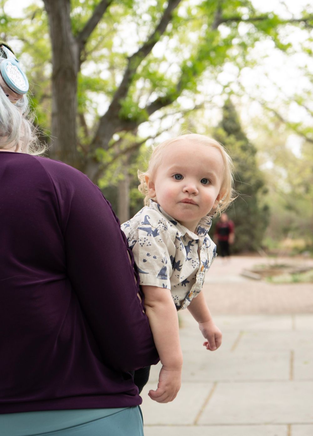 A baby in a floral shirt looks over an adult's shoulder outdoors near trees.