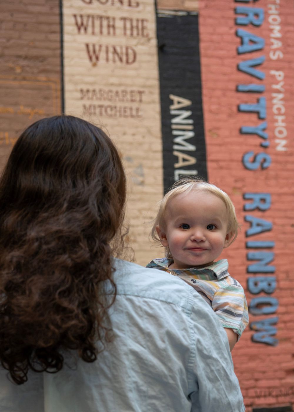 A woman holds a smiling baby in front of a brick wall painted with book titles including Gone with the Wind and Gravity's Rainbow.