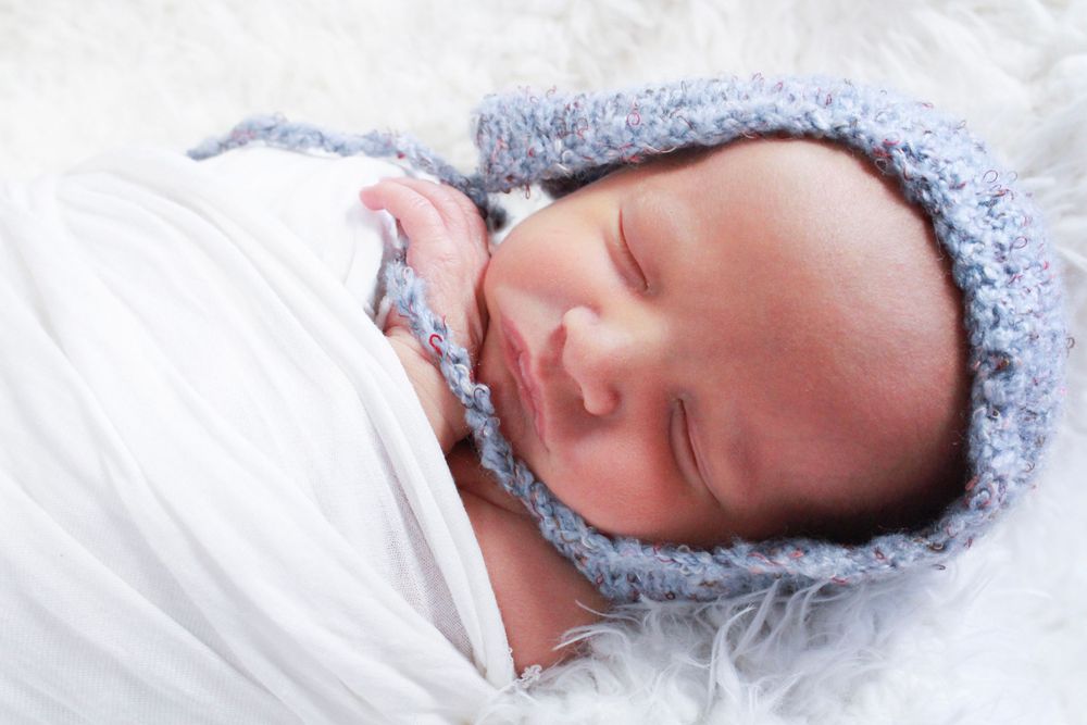 Sleeping newborn wrapped in white blanket wearing soft blue bonnet.