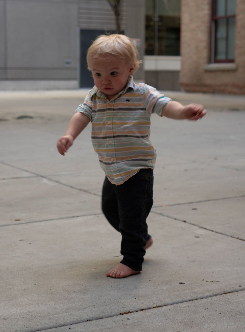 Barefoot toddler boy in striped shirt and dark pants walking outdoors on a concrete sidewalk near a building.