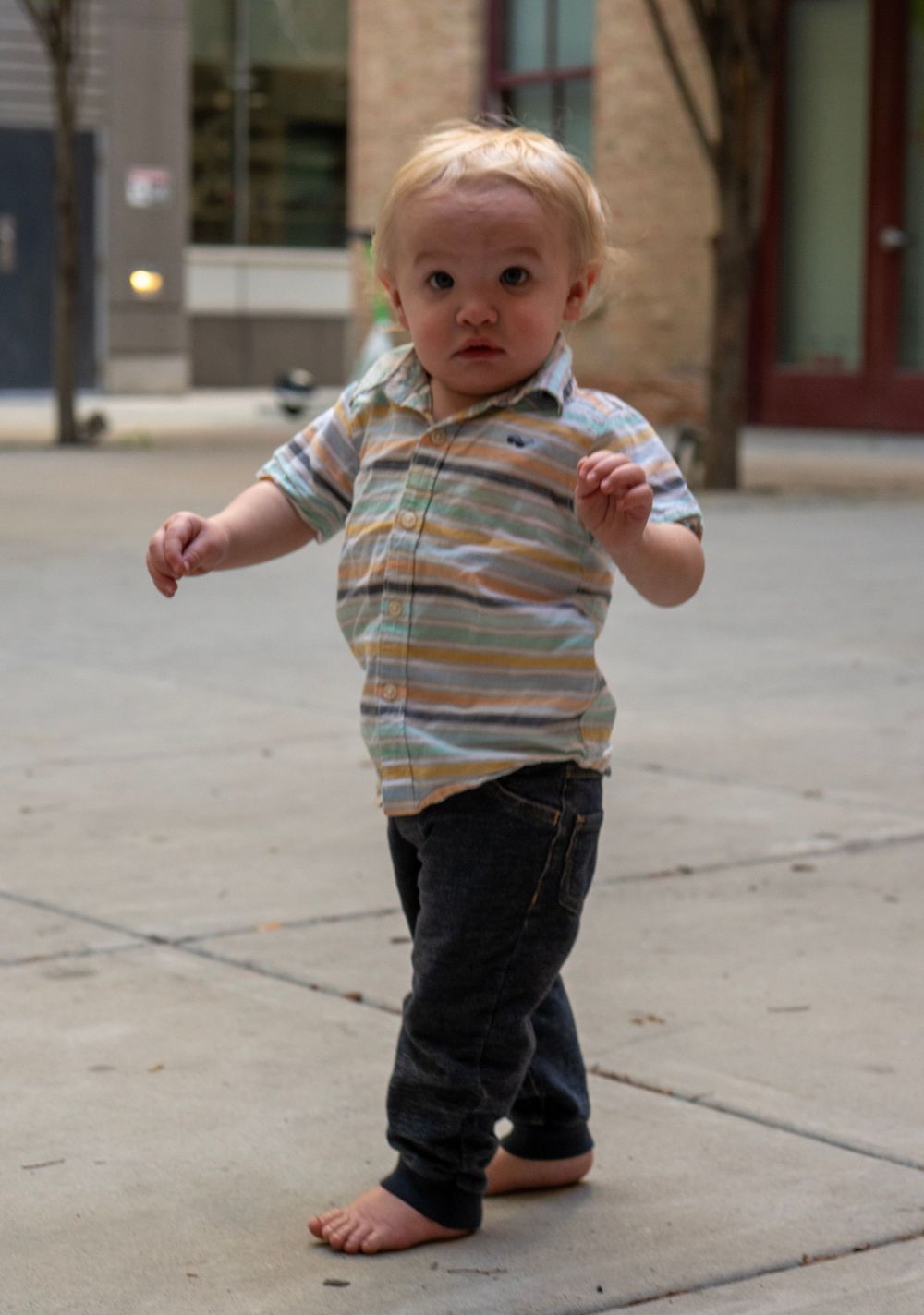 Toddler boy in striped polo shirt and jeans walking barefoot on a city sidewalk.