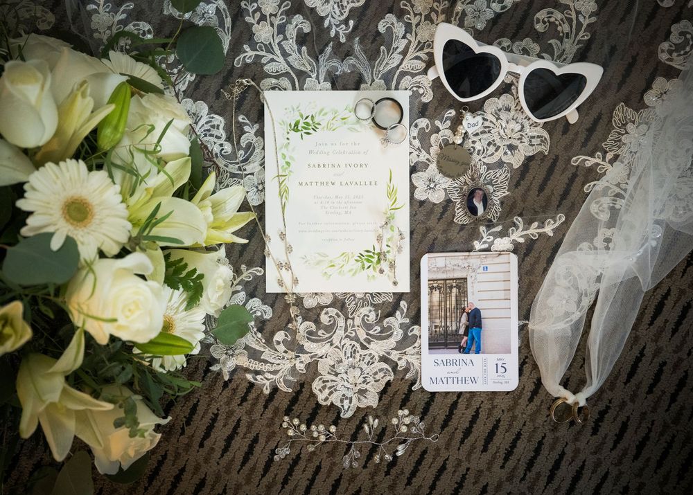 Wedding invitation suite laid out with heart-shaped sunglasses, white bouquet and vintage lace on wooden background.