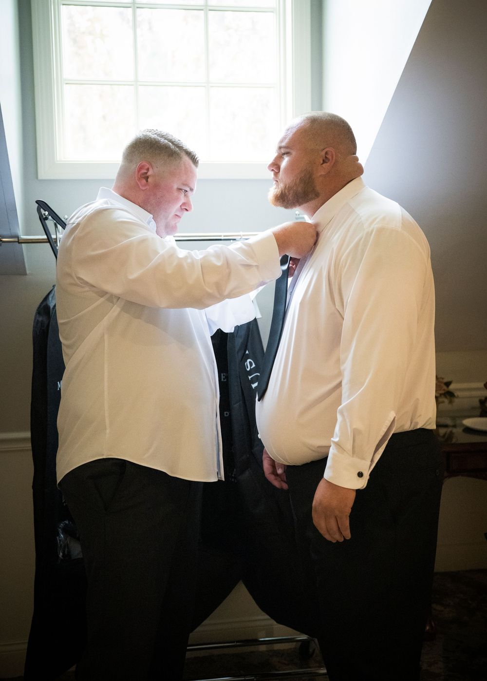 Two people in white dress shirts help each other adjust formal attire while getting ready in a sunlit room.