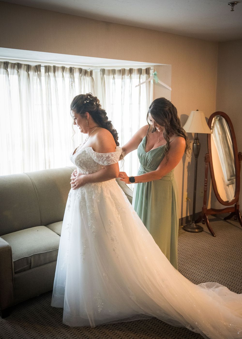 Bridesmaid in mint green dress helps adjust the bride's flowing white wedding gown in a hotel room.