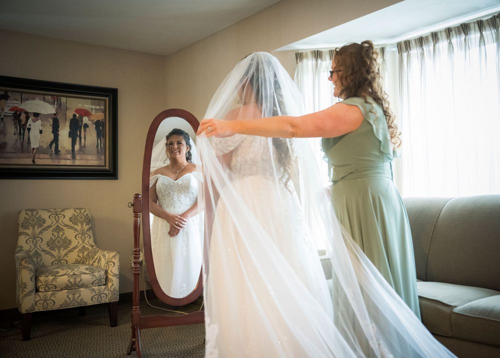 A figure in a flowing wedding gown stands before an ornate mirror while someone adjusts their veil.