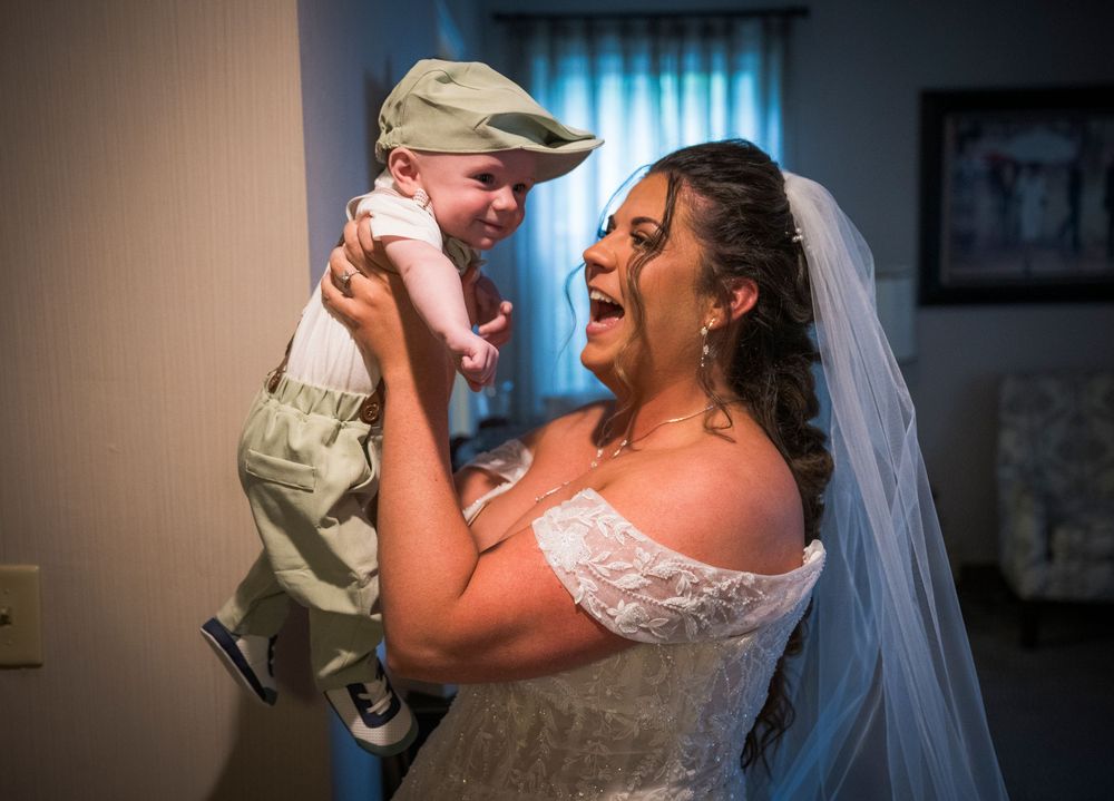 A bride in a white dress joyfully lifts a small child wearing a cap during a wedding celebration.
