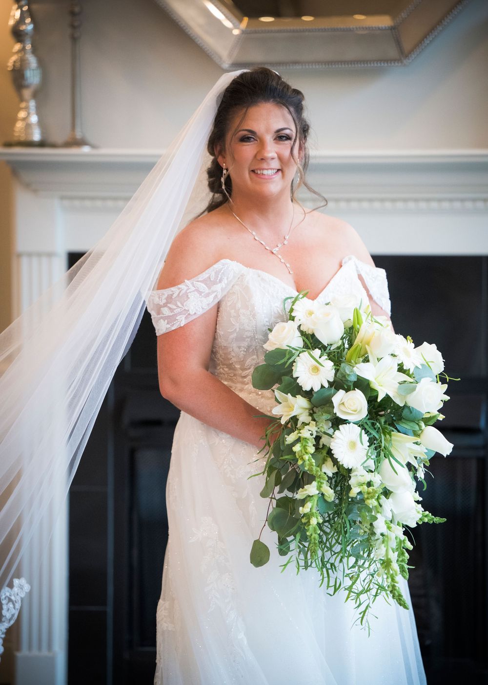 Beautiful bridal portrait featuring an off-shoulder lace wedding gown and cascading white floral bouquet by a fireplace mantle.