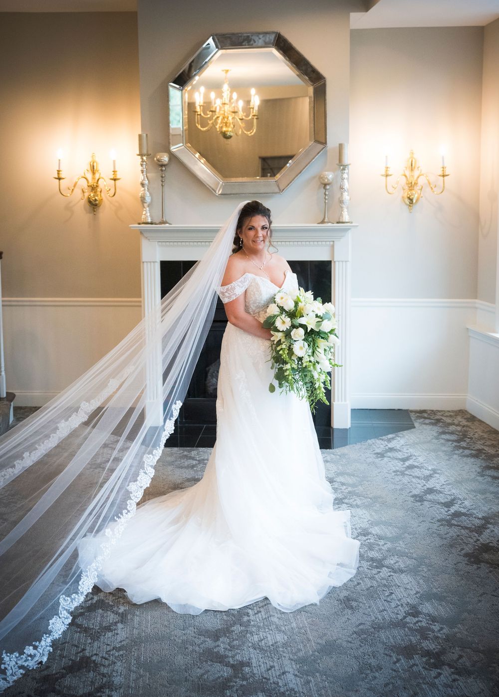 An elegant white wedding gown and flowing veil photographed in front of a fireplace with ornate gold sconces and mirrors.