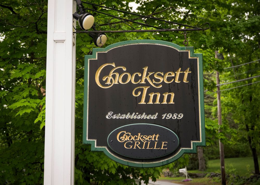 A classic black and gold wooden sign for Crocksett Inn and Crocksett Grille, established in 1989, hangs outdoors among green trees.