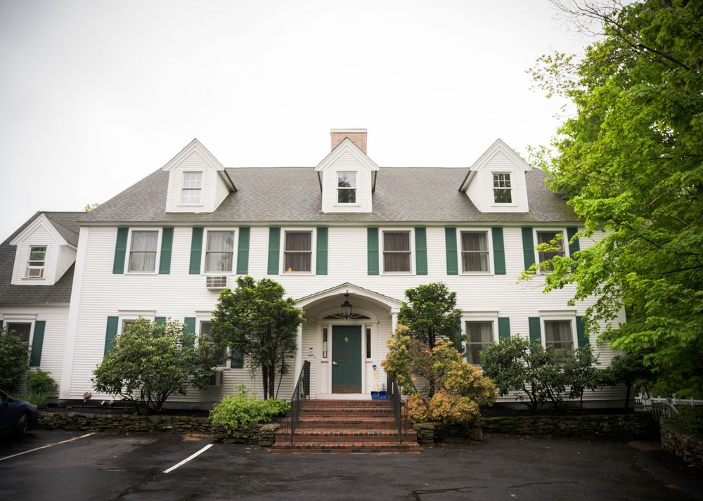 Large white colonial style house with green shutters, dormers, and a grand entrance portico set among mature trees.