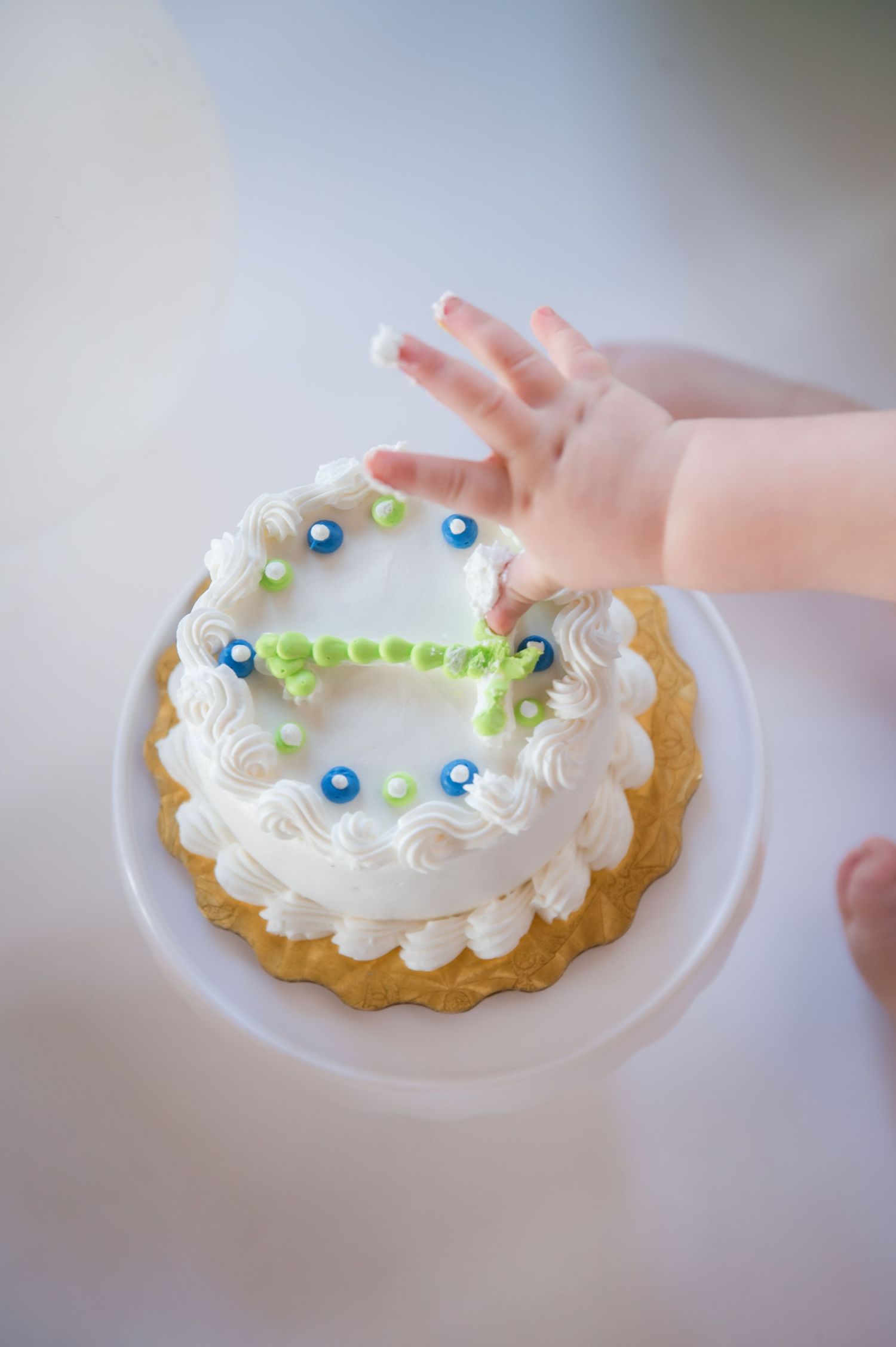 A baby's small hand reaches for a white frosted birthday cake decorated with blue dots and green accents on a pedestal stand.