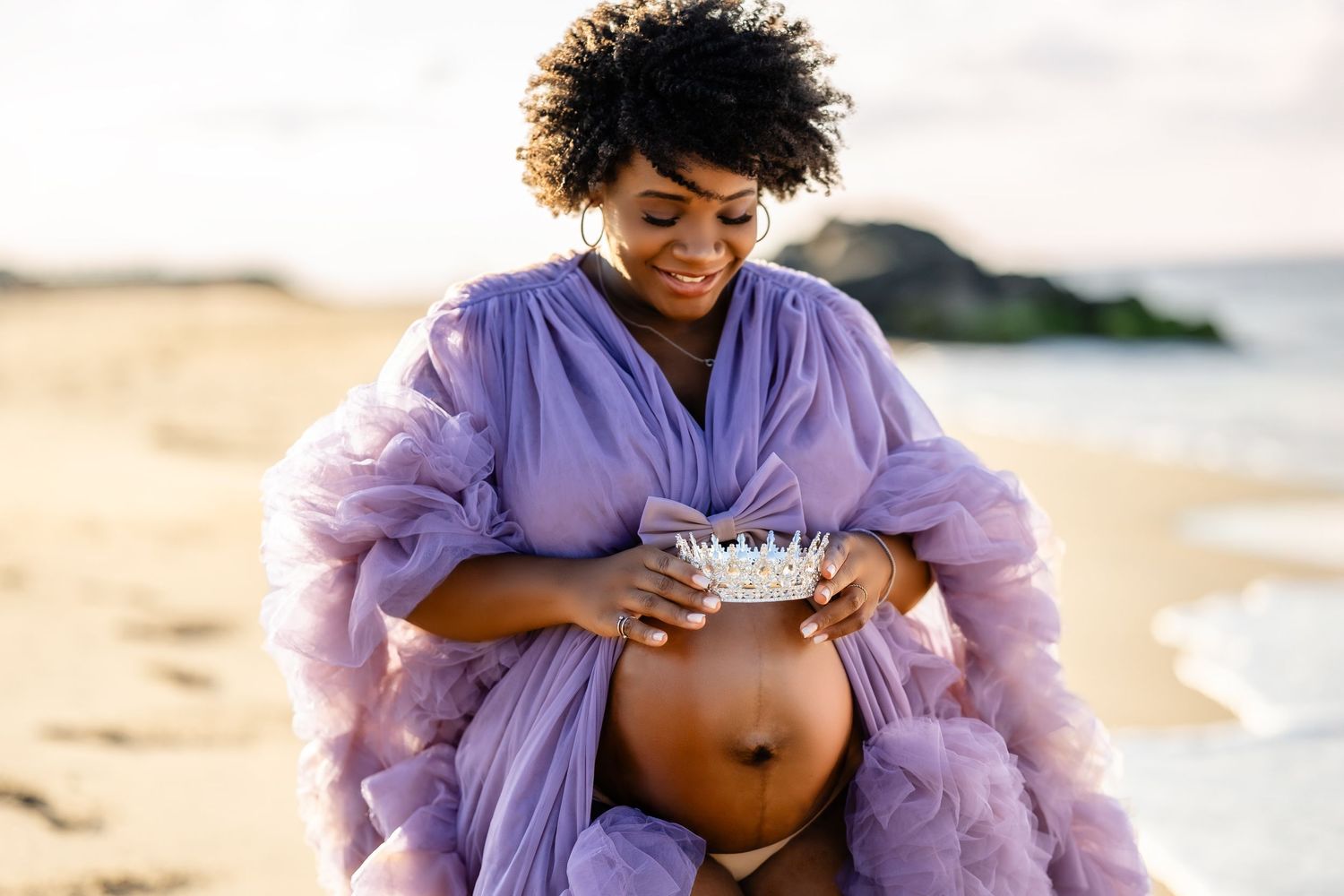A pregnant woman in a flowing pink dress poses at sunset among large rocks during a maternity photoshoot.