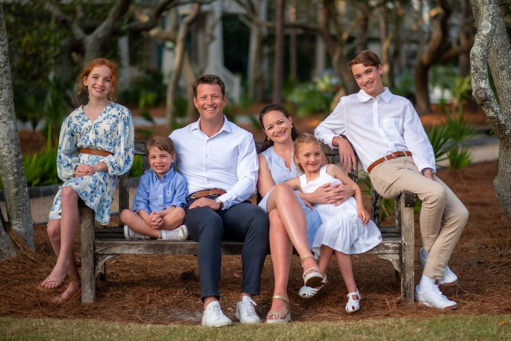 Family sitting together outdoors on a rustic wooden bench surrounded by trees in casual summer attire.