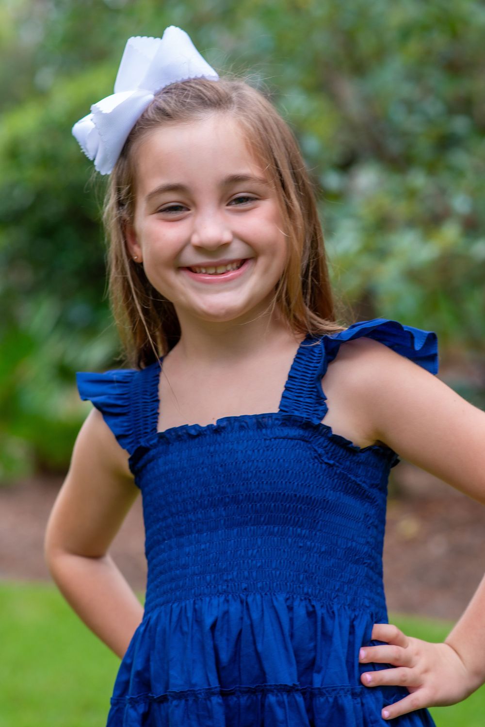 Child wearing blue ruffled summer dress and white hair bow poses outdoors with a bright smile and hands on hips.