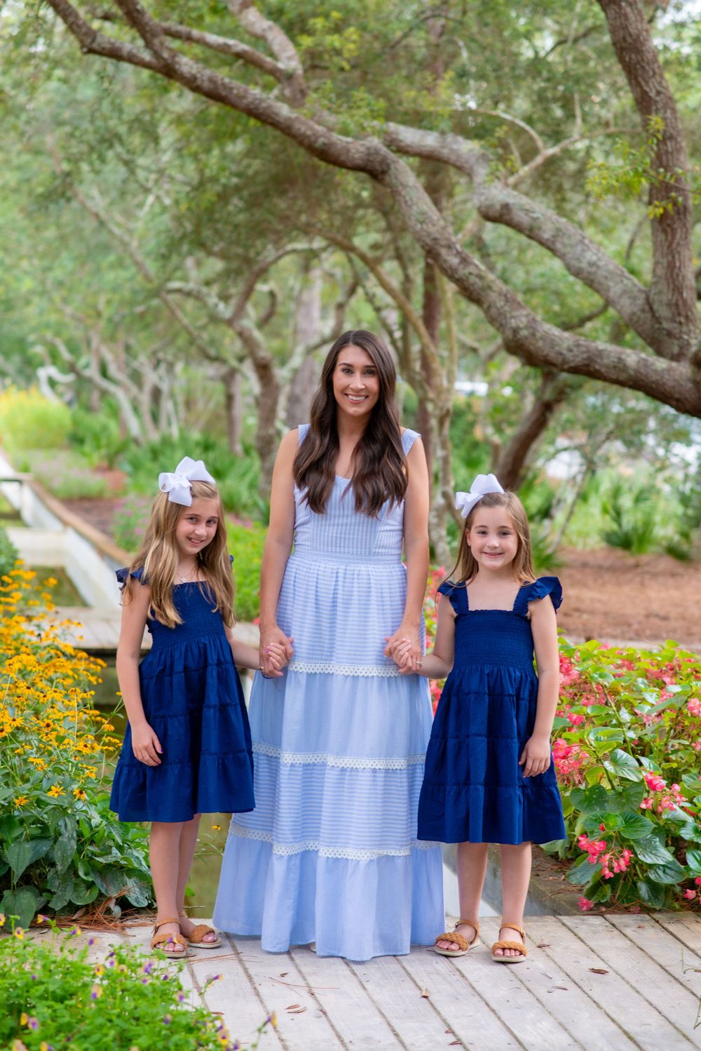 Three people wearing coordinating blue dresses pose together on a garden path surrounded by colorful flowers and trees.