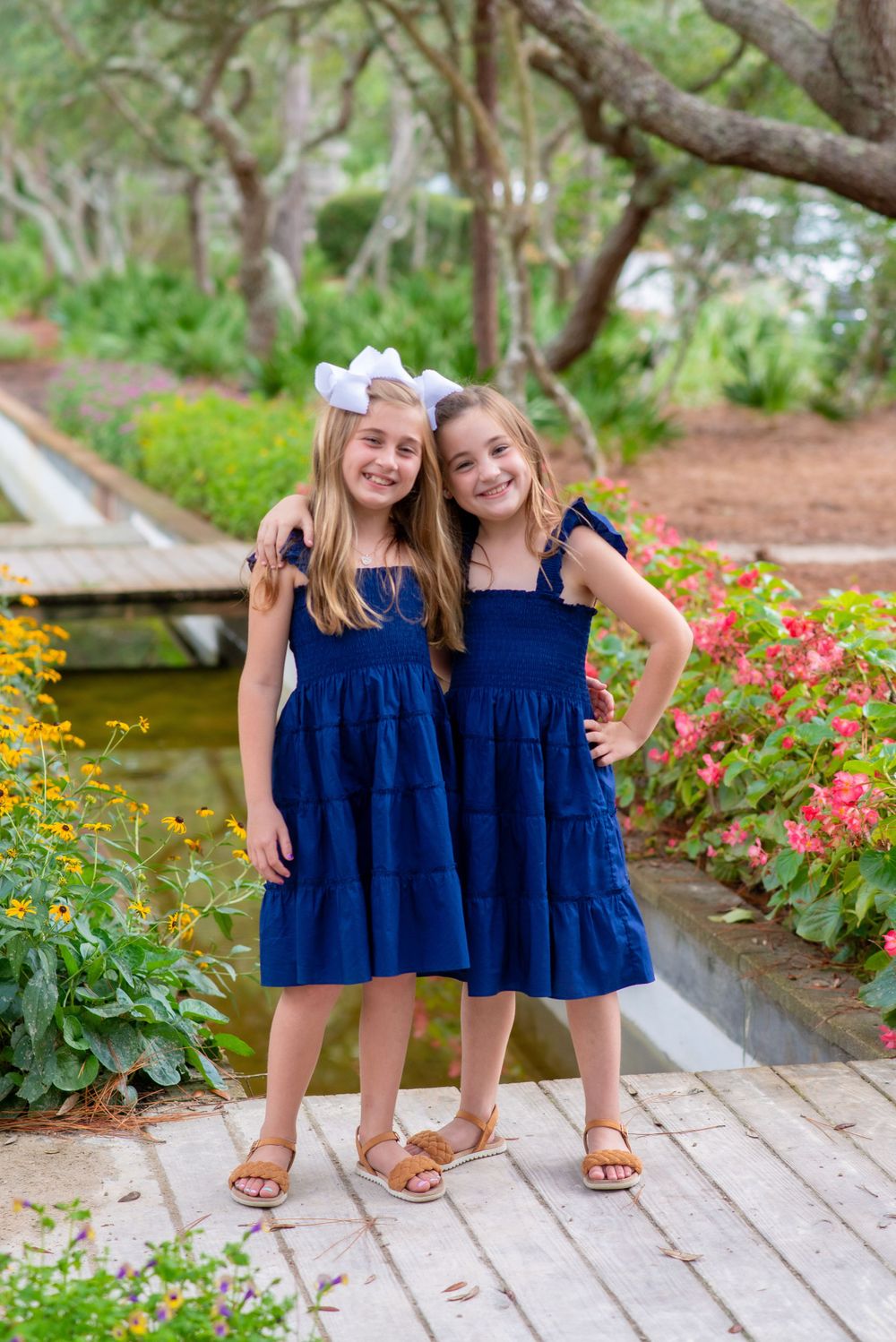 Two young girls in matching navy blue summer dresses pose together in a garden setting with flowers and greenery.