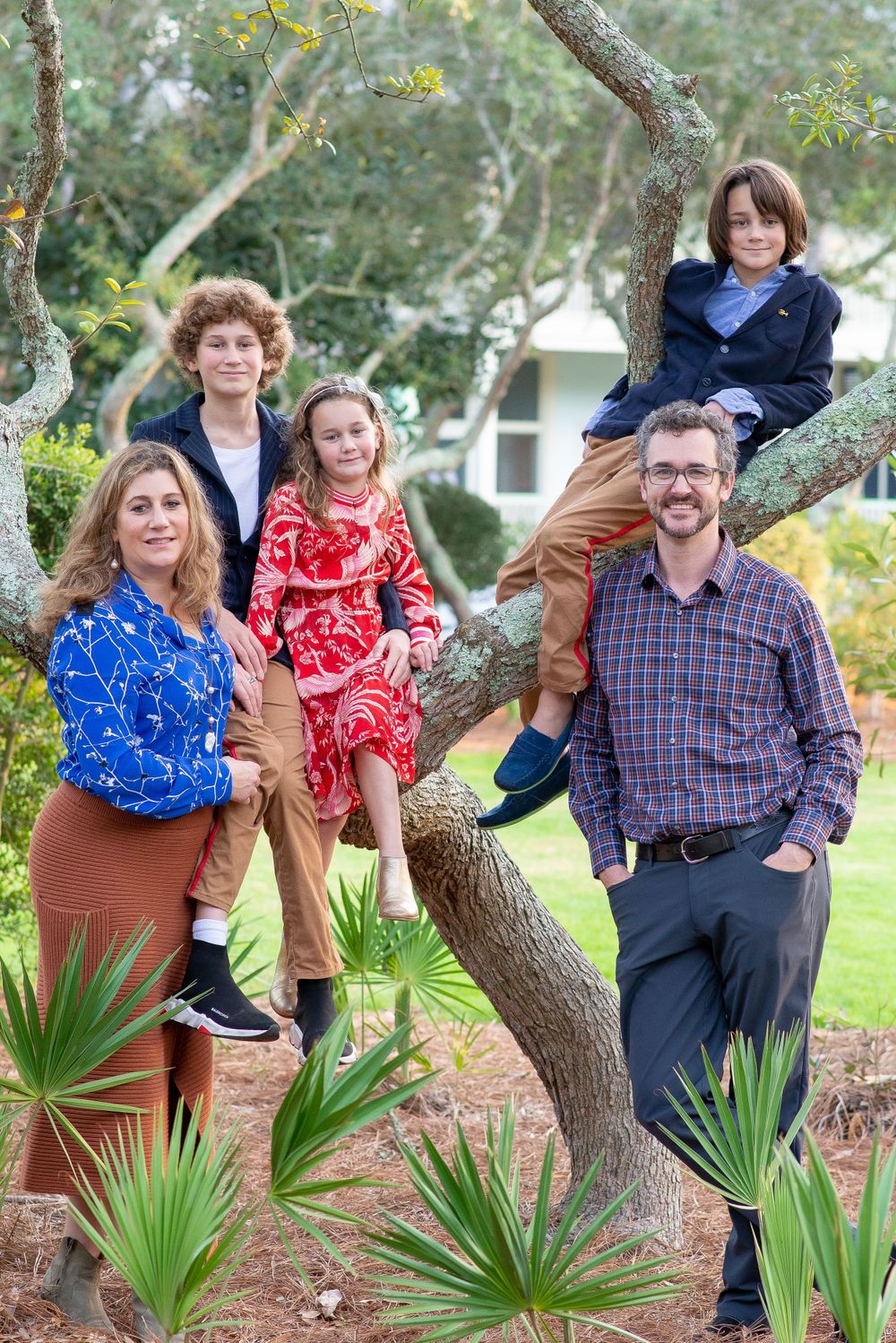 A family poses together around a tree in an outdoor setting with palm plants, wearing colorful casual attire.