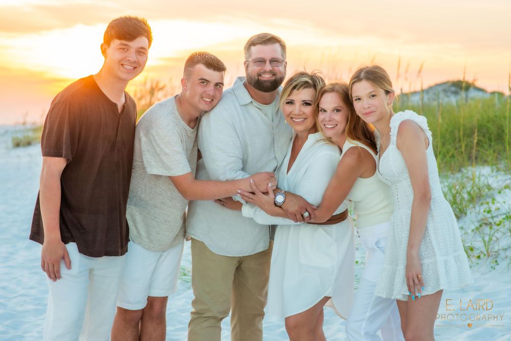 A group of people in light colored clothing posing together on a beach during golden sunset hour.