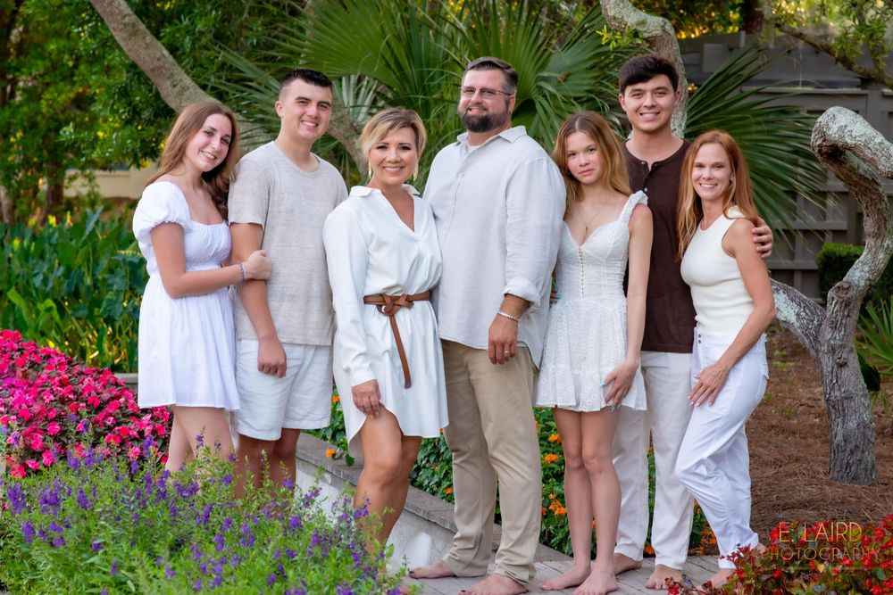 A family wearing white outfits poses together in a beautiful garden with colorful flowers and greenery.