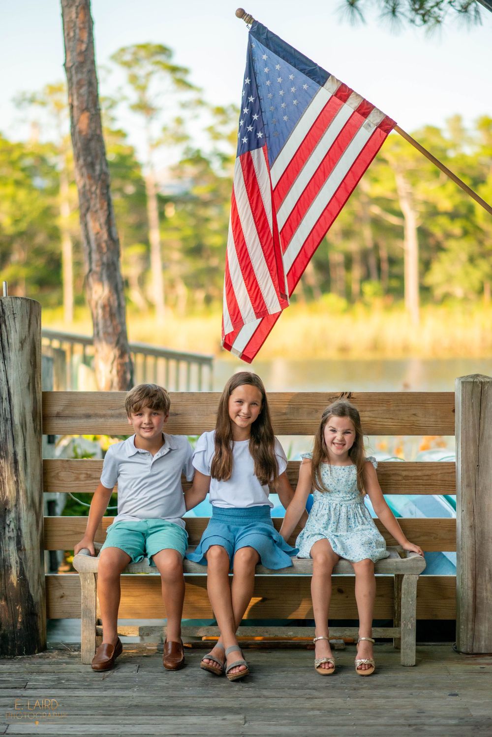 Three children sit on a wooden bench under an American flag on a dock with pine trees in the background.