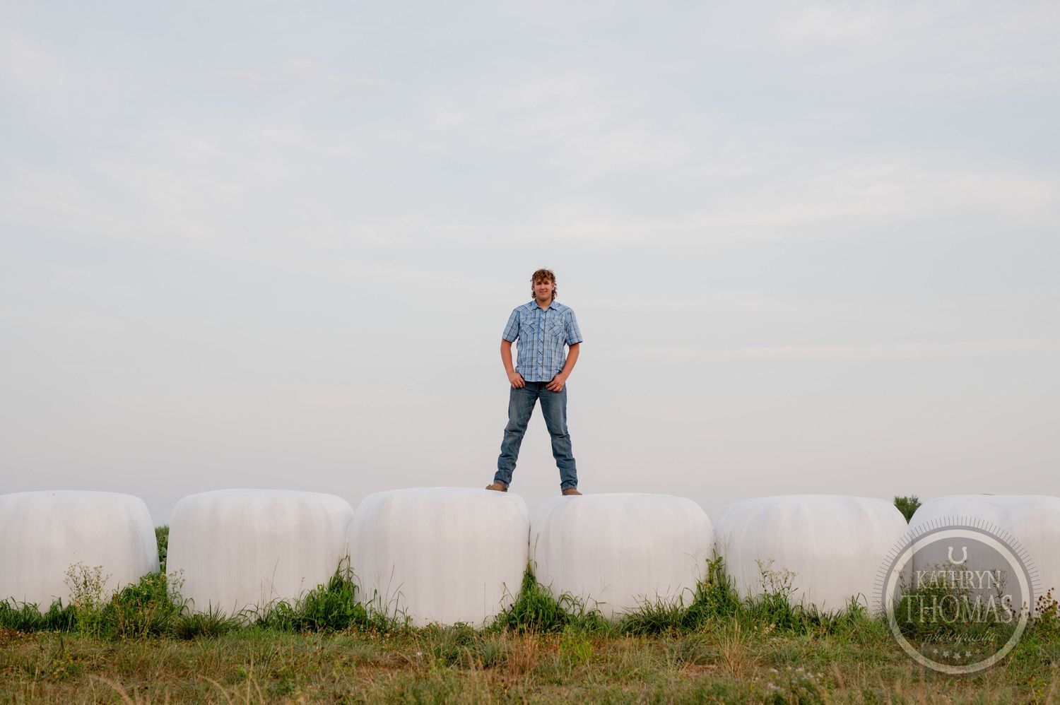A person stands on white hay bales against a bright sky in a rural setting.
