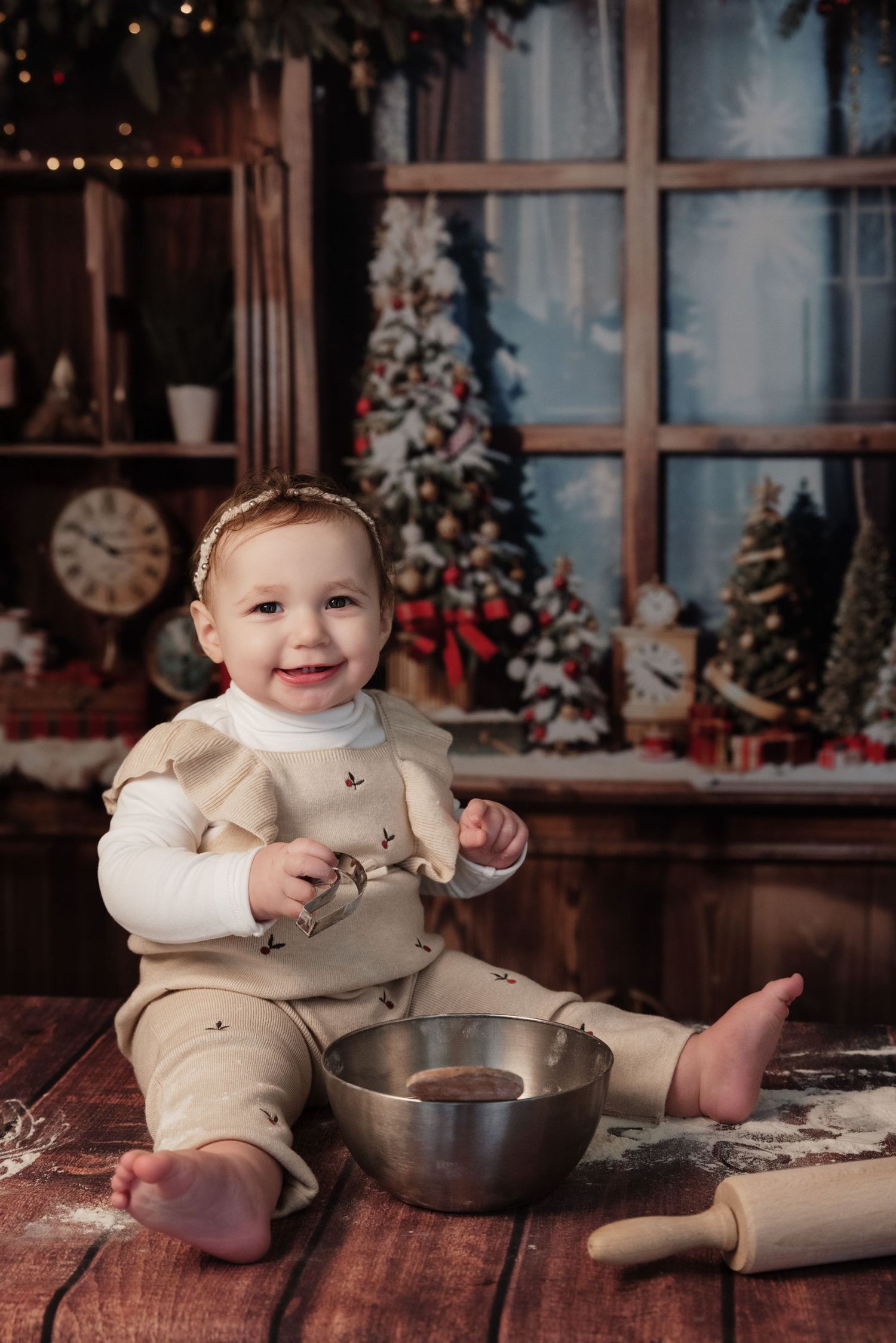 A small child in a cream-colored outfit sits on wooden floor with a mixing bowl and rolling pin near a festive Christmas scene.