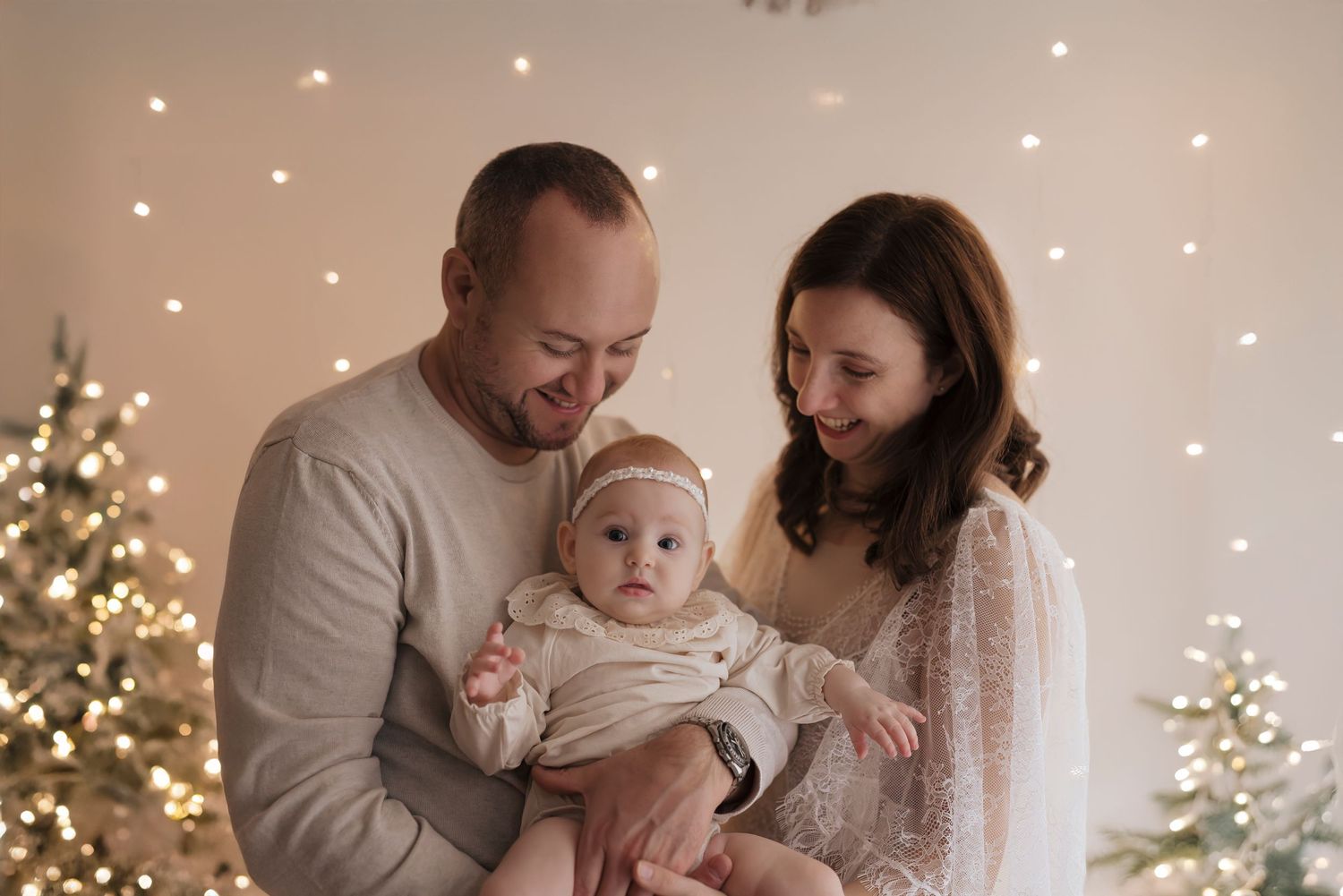 Family sharing a tender holiday moment with their baby in front of illuminated Christmas trees and twinkle lights.
