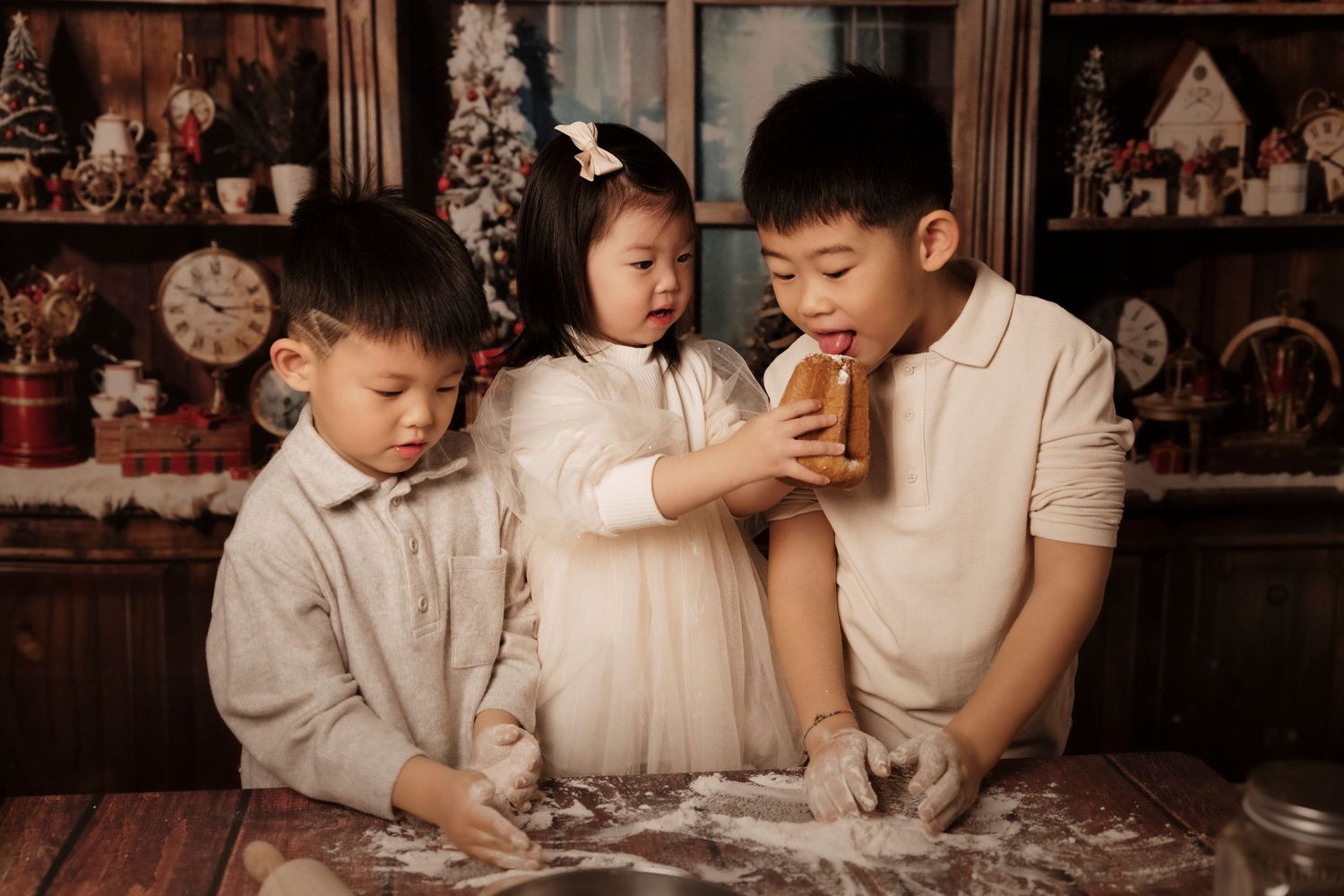 Three children in light colored clothing working together with flour and dough on a wooden table in a cozy kitchen.