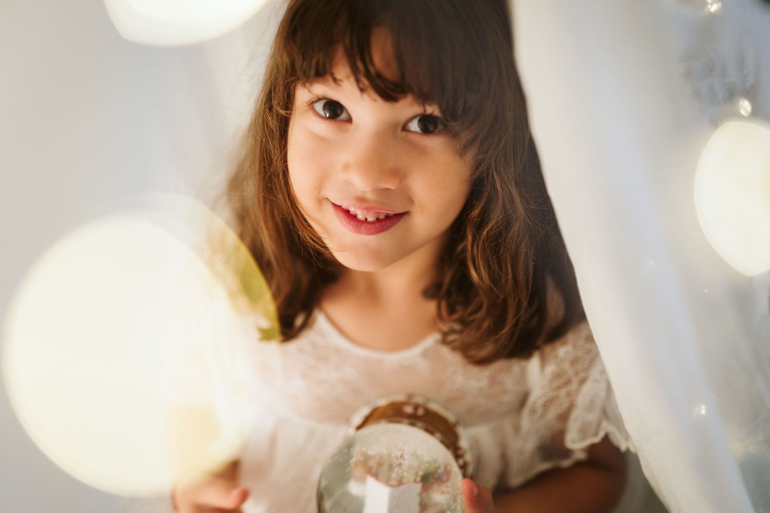 A child holding a sparkly glass orb smiles while soft bokeh lights create a dreamy atmosphere in the background.