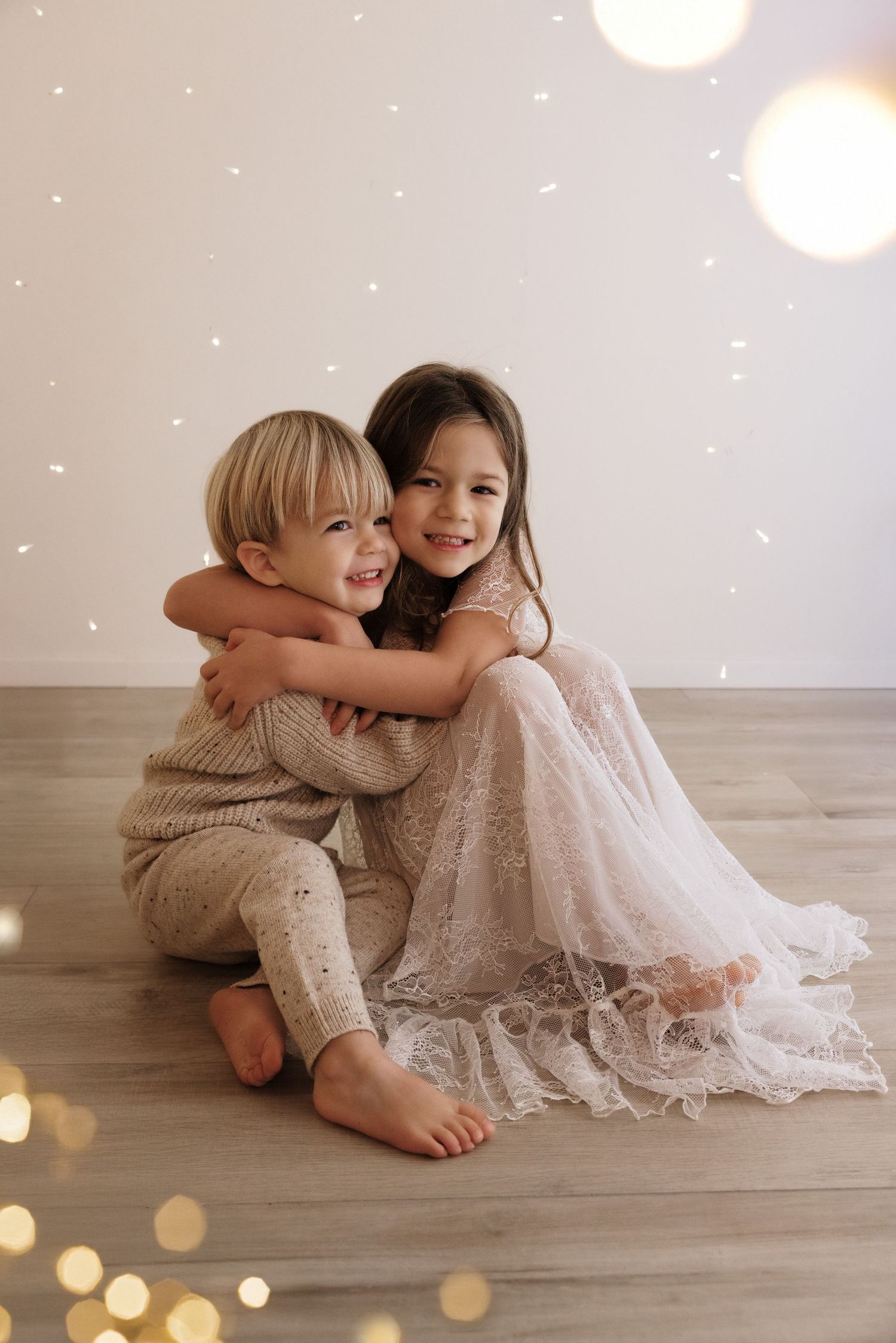 Two children embrace while sitting on hardwood floor with glowing lights and sparkles in a dreamy indoor setting.