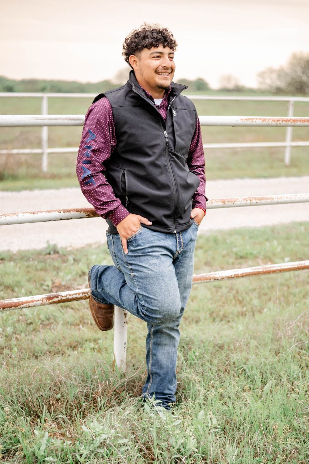 Person in a black vest and plaid shirt leans casually against a wooden fence post in a rural setting.