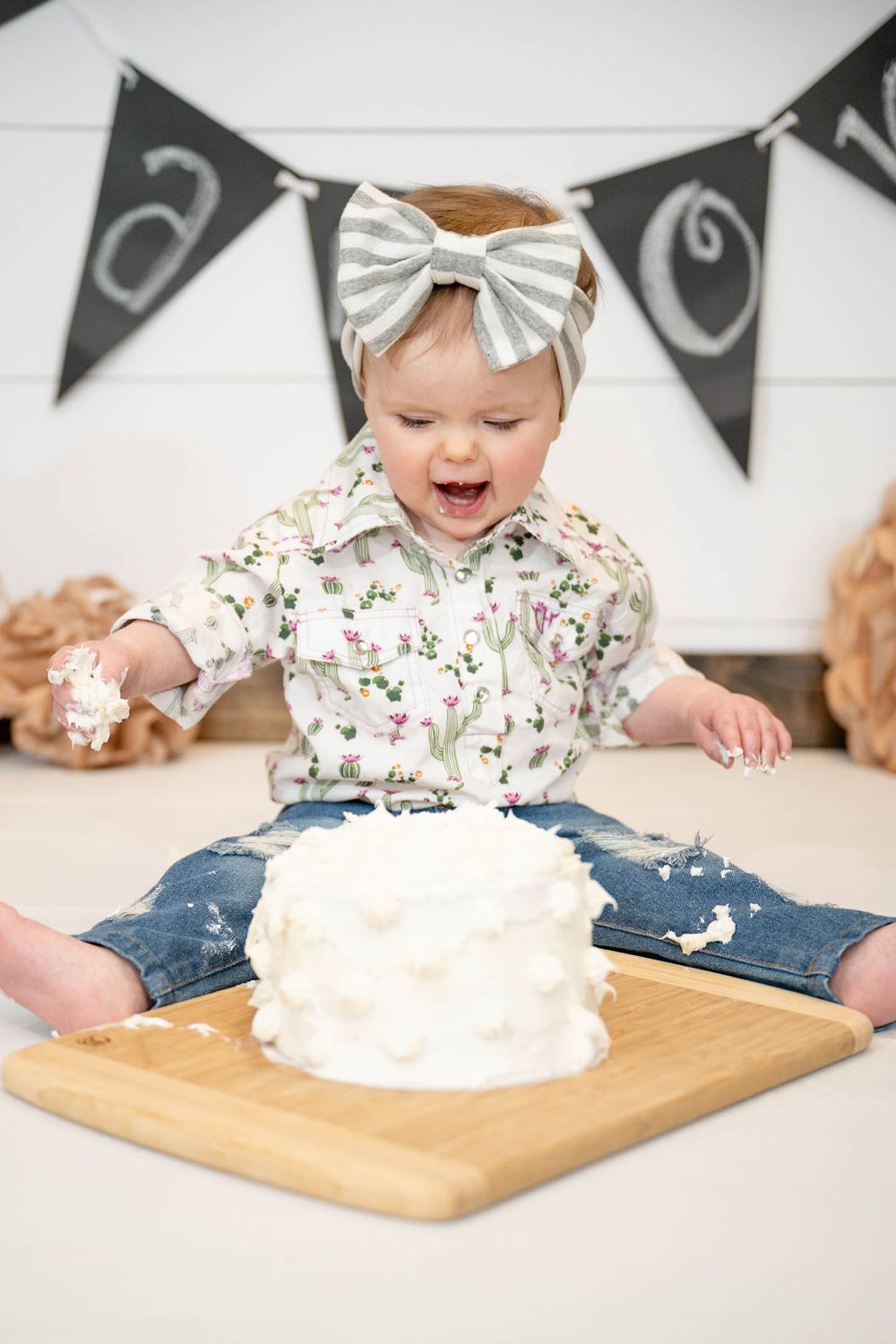 Baby in floral shirt and silver bow celebrates with white cake on wooden board beneath black and white banner.