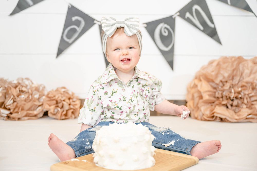 First birthday celebration scene with a white cake, grey and white pennant banner, and brown tissue paper decorations.