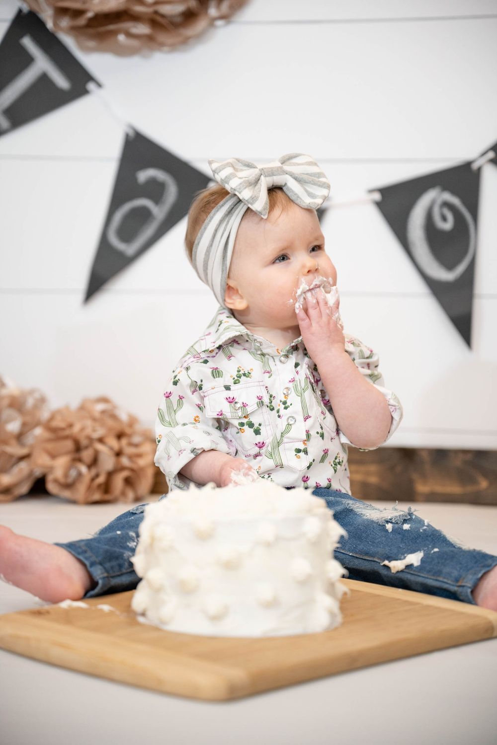 A small child wearing a floral outfit and bow headband tastes frosting while sitting with a white cake on a wooden board.