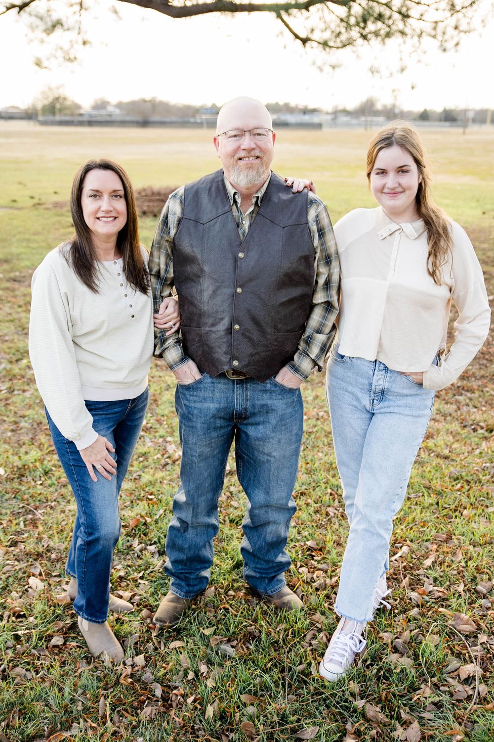 Three people dressed in casual sweaters and jeans pose outdoors on a sunny autumn day in a grassy field.