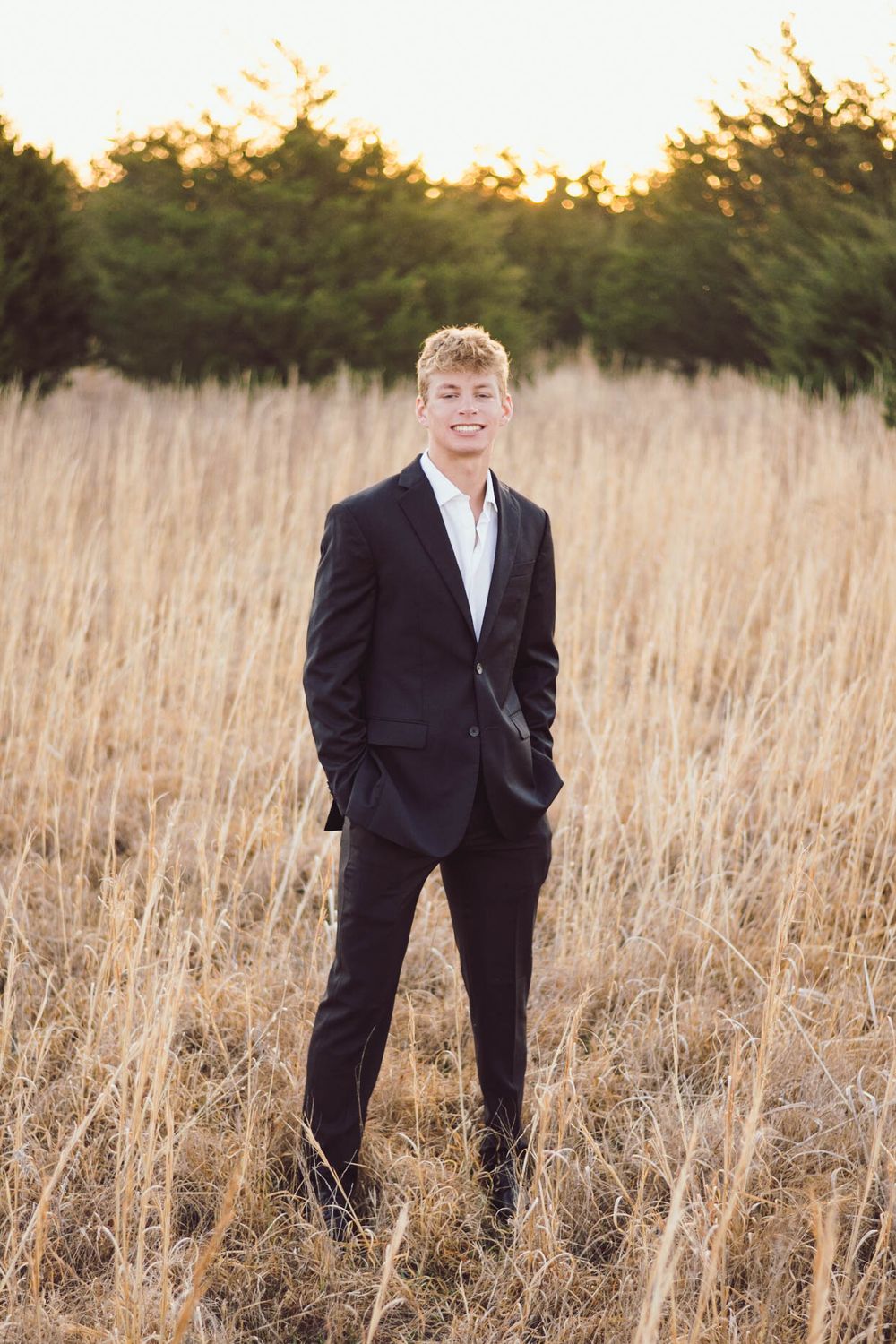 Young person in formal black suit stands in tall dry grass field at sunset.