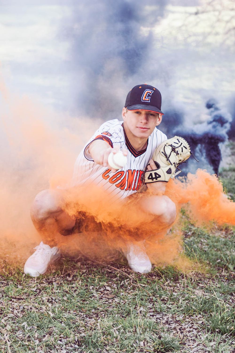 Baseball player crouches in catcher's stance surrounded by dramatic orange smoke on a grassy field.