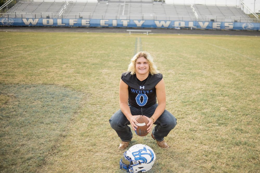 Football player sits on a dry grass field wearing a black t-shirt with helmet and stadium bleachers in background.