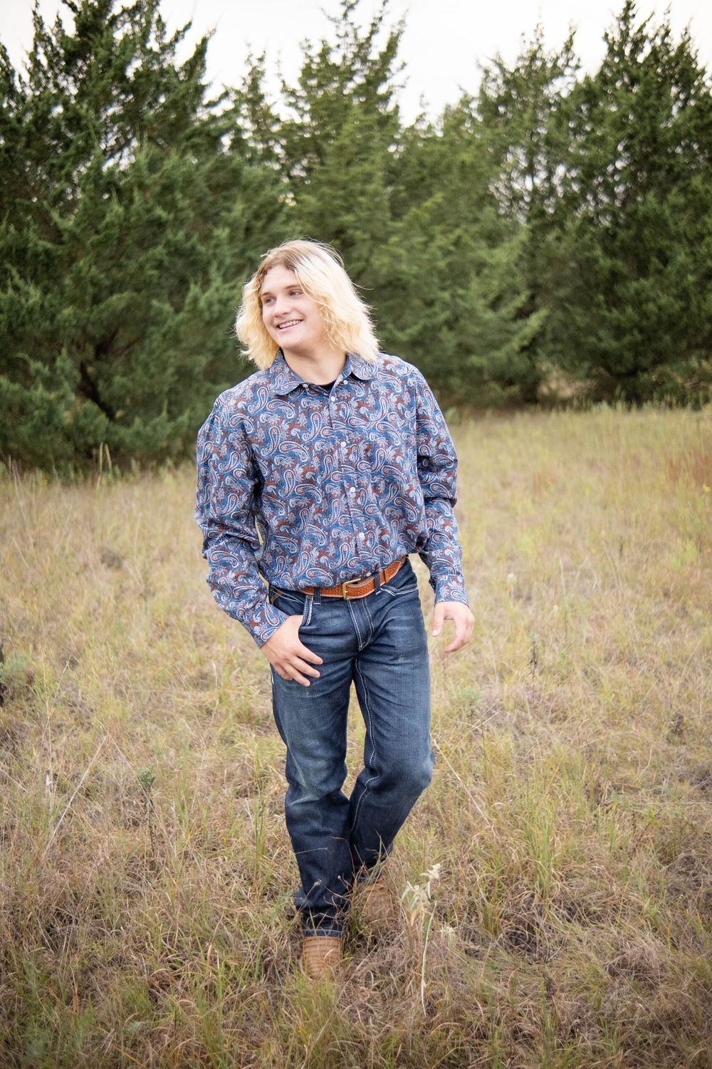 Someone wearing a blue floral shirt and jeans stands in a field with evergreen trees in the background.