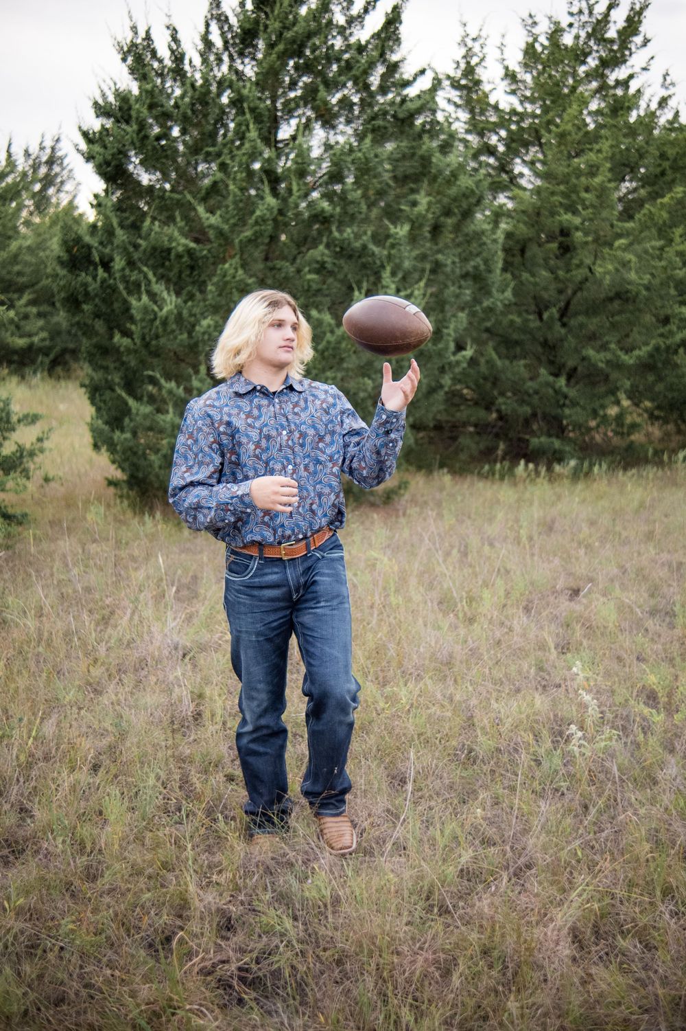 A casual figure holds a football while standing in a grassy field with evergreen trees in the background.