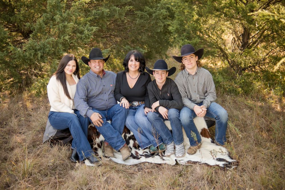 A casual outdoor family photo of five people in blue jeans sitting together on a blanket against a natural backdrop.