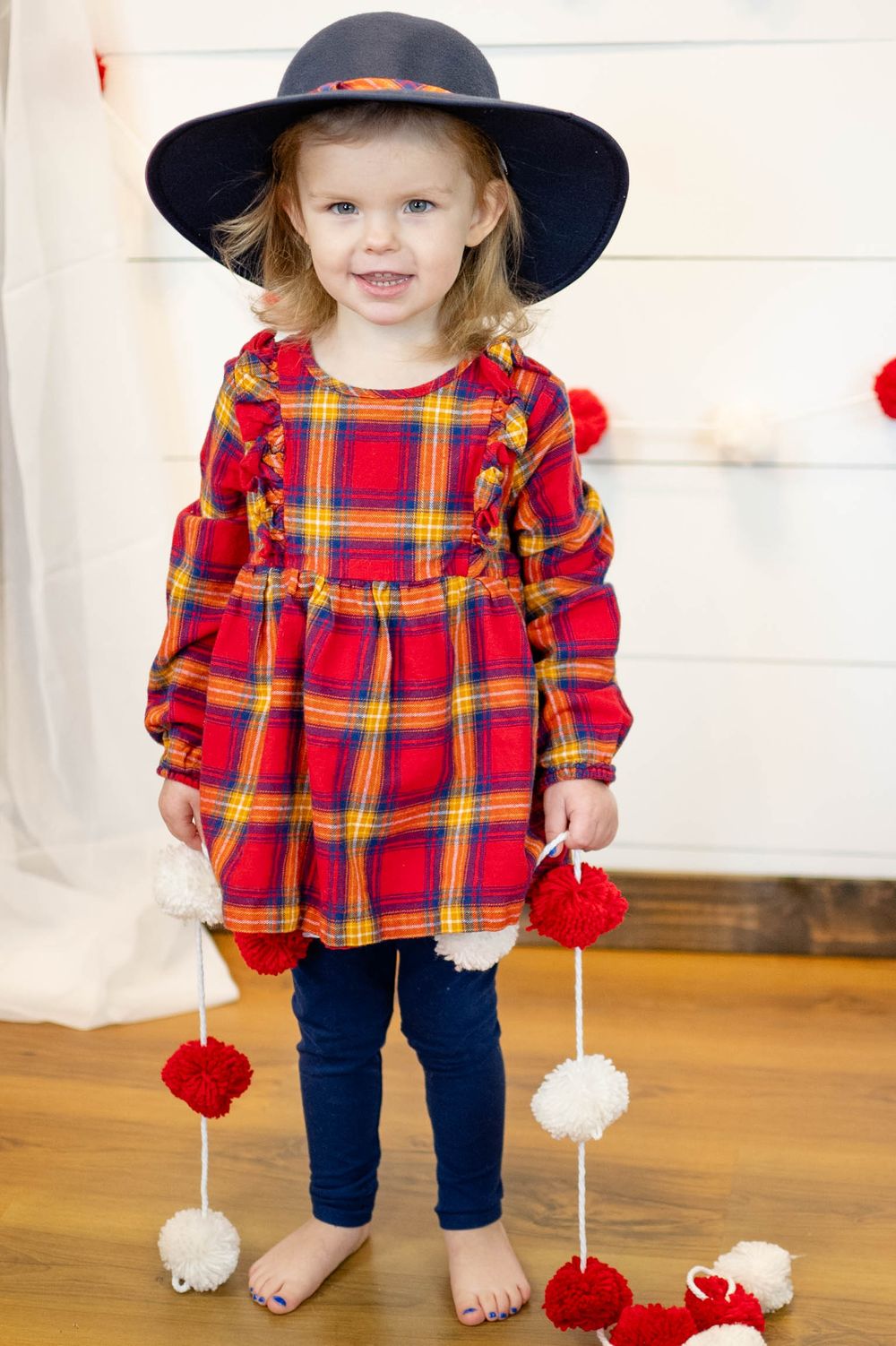 A child wearing a red plaid dress, navy hat, and blue leggings holds decorative red and white pom pom garland.