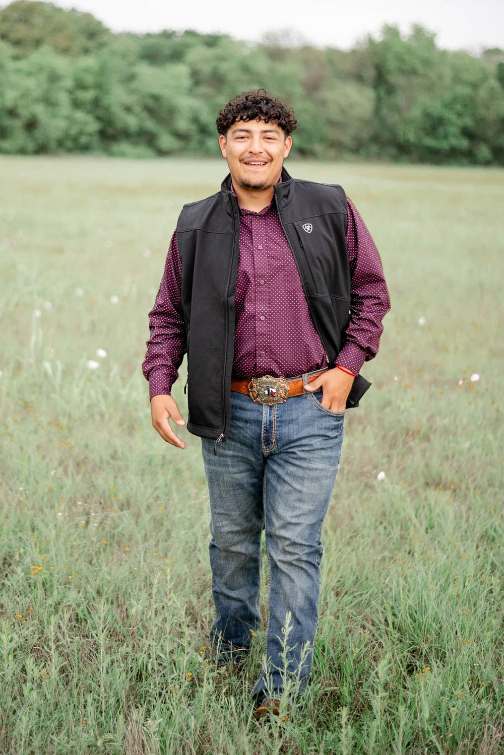 Someone stands in a grassy meadow wearing a black vest, burgundy button-down shirt and blue jeans on a sunny day.