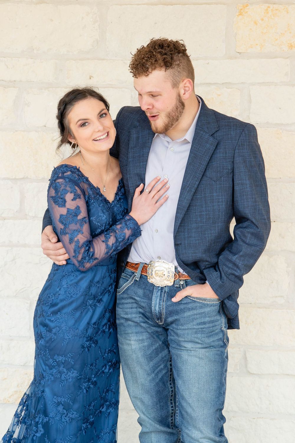 A couple in matching blue outfits pose together against a light stone wall during a romantic photoshoot.
