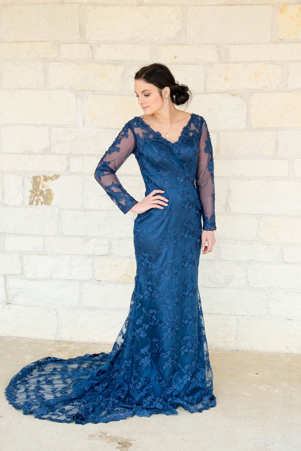 An elegant navy blue lace gown with long sleeves and a dramatic train is showcased against a white brick wall.