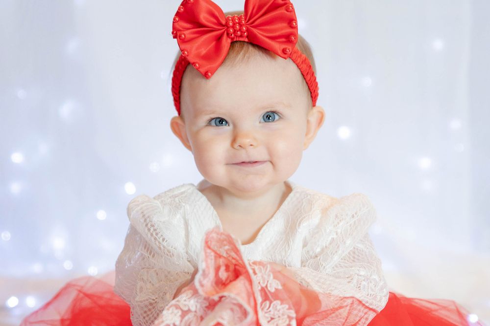 Baby in white lace dress and red bow headband poses against sparkly background.