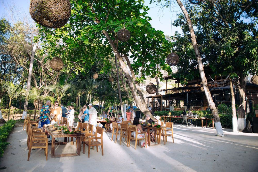 Outdoor restaurant seating area with wooden chairs and woven lanterns hanging from trees in a tropical garden setting.